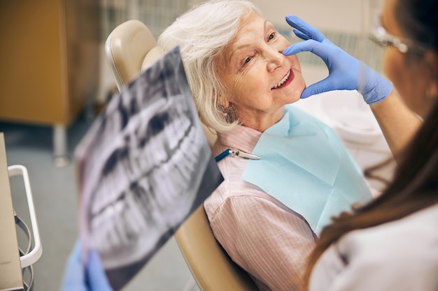 Portrait of smiling female patient sitting on the dental chair while woman doctor touching face of client