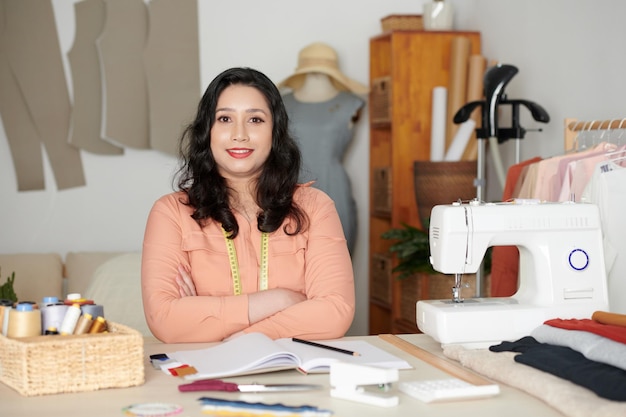 Portrait of smiling female tailor sitting at table with sewing machine basket of threads fabrics and...