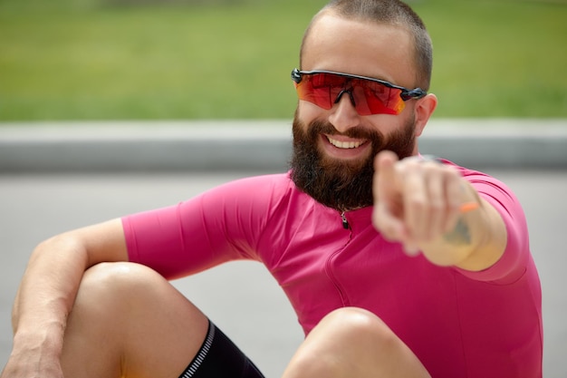 Portrait of a smiling fitness man resting while sitting on a ground at the track field outdoors pointing his finger at the camera