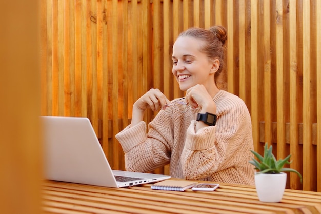 Portrait of smiling friendly pleased beautiful woman with bun hairstyle wearing beige sweater working on laptop against wooden wall looking at notebook screen and holding optical eyeglasses in hands