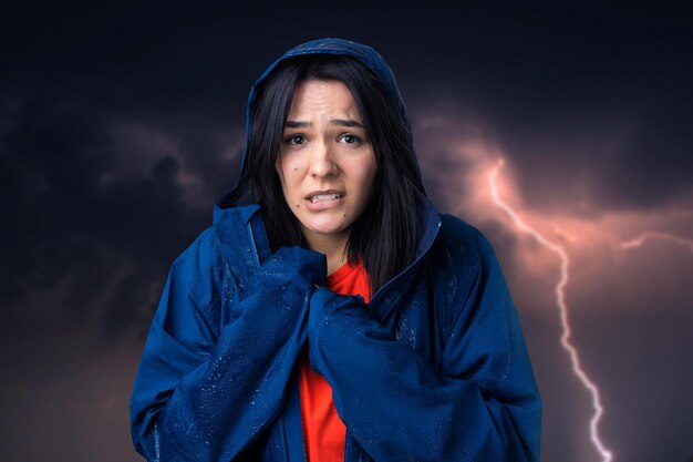 Portrait of a smiling girl dressed in blue raincoat in drops posing with hood against the background of a gloomy sky with lightning