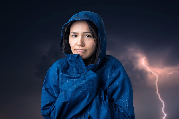 Portrait of a smiling girl dressed in blue raincoat in drops posing with hood against the background of a gloomy sky with lightning