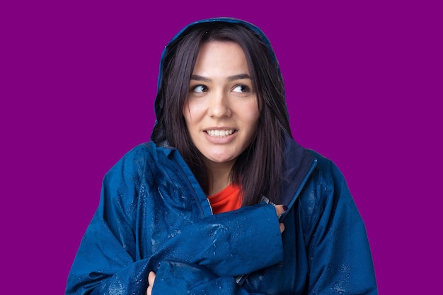 Portrait of a smiling girl dressed in blue raincoat in drops posing with hood on grey background in a studio