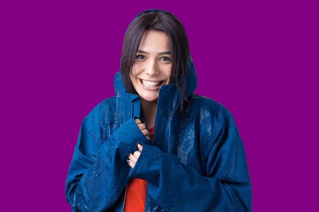 Portrait of a smiling girl dressed in blue raincoat in drops posing with hood on grey background in a studio