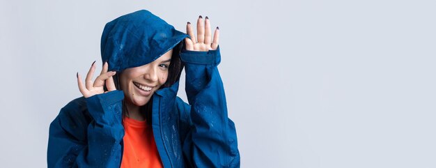 Portrait of a smiling girl dressed in blue raincoat in drops posing with hood on grey background in a studio