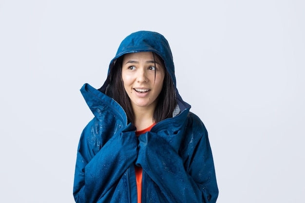 Portrait of a smiling girl dressed in blue raincoat in drops posing with hood on grey background