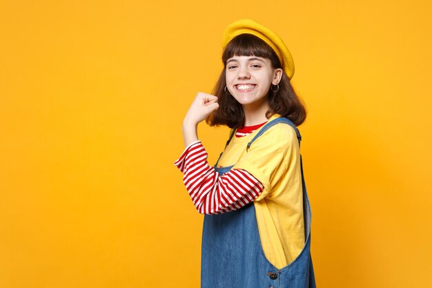 Portrait of smiling girl teenager in french beret, denim sundress showing biceps, muscles isolated on yellow wall background in studio. People sincere emotions, lifestyle concept. Mock up copy space.