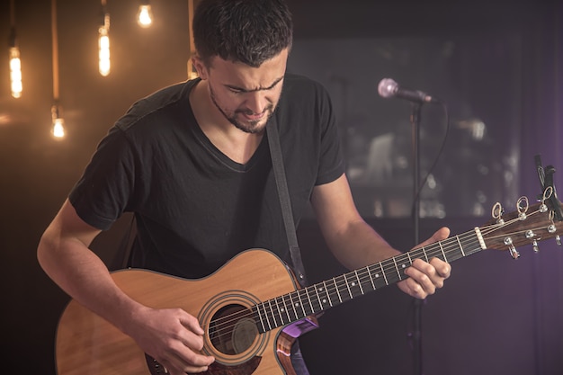 Portrait of smiling guitarist in black t-shirt playing acoustic guitar on blurred studio dark space.