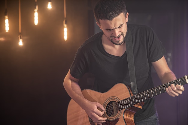 Portrait of smiling guitarist in black t-shirt playing acoustic guitar on blurred studio dark wall.