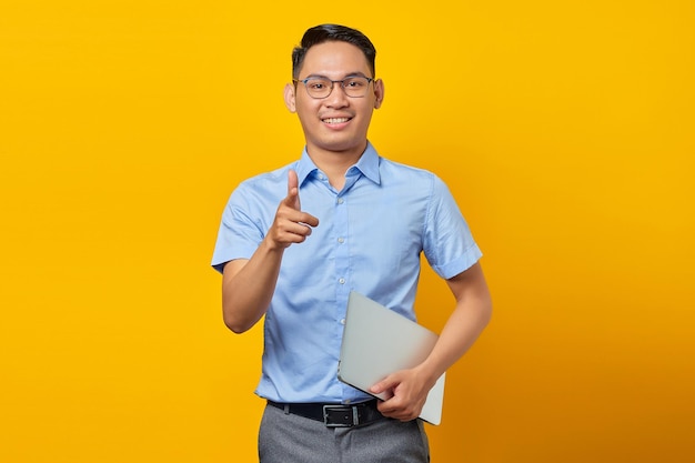 Portrait of smiling handsome Asian man in glasses holding laptop and pointing fingers at camera isolated on yellow background businessman and entrepreneur concept