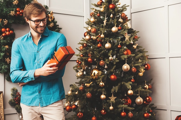 Portrait of smiling handsome man holding gift. Sexy bearded male posing near Christmas tree with present. 