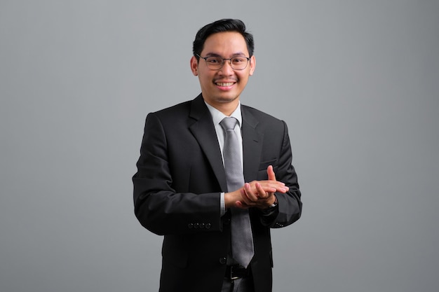 Portrait of smiling handsome young businessman in formal suit clapping hands and looking at camera isolated on grey background