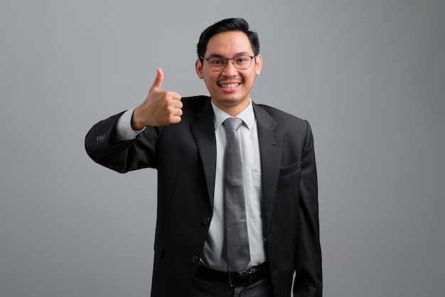 Portrait of smiling handsome young businessman in formal suit showing thumb up isolated on grey background