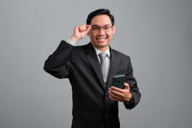 Portrait of smiling handsome young businessman in formal suit using smartphone and adjusting eyeglasses looking at camera isolated on grey background