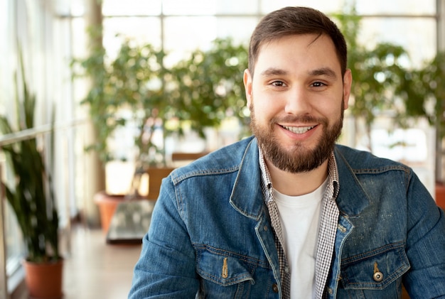 Portrait smiling handsome young man with beard in modern corporate environment near window. Smart man in office in casual wear, jean jacket. Attractive businessman freelancer smiling in cafe.