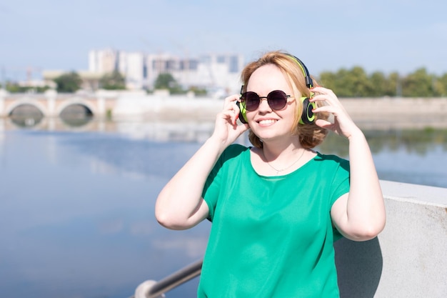 Portrait of a smiling happy girl in sunglasses and headphones listening to music on the riverbank