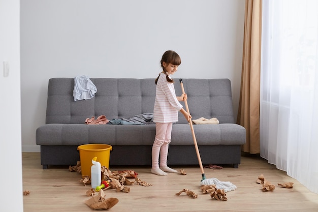 Portrait of smiling happy little girl washing the floor with a mop at home cleaning living room standing in mess helping mother with household chores