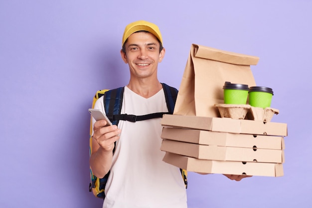 Portrait of smiling happy man courier in yellow cap and Tshirt holding food order pizza boxes and coffee to go using cell phone looking at camera isolated on purple background