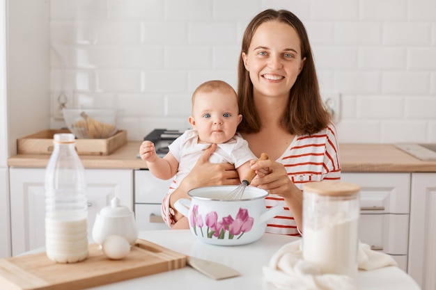 Portrait of smiling happy woman wearing striped shirt sitting at table with her baby daughter in kitchen mixing dough for homemade pastry together looking at camera