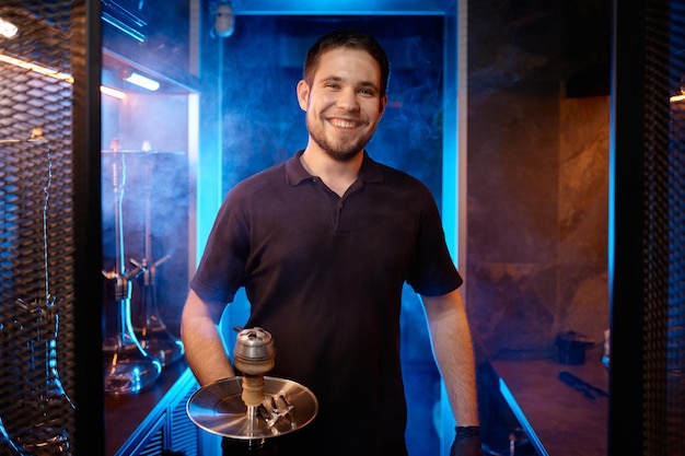 Portrait of smiling hookah man holding traditional eastern accessory for smoking. Guy carries shisha to client