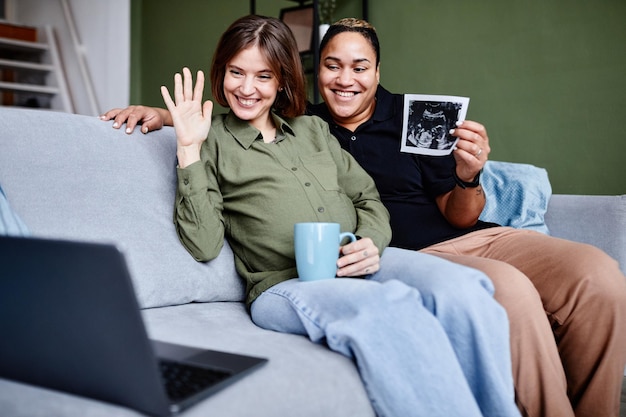 Portrait of smiling lesbian couple expecting baby and showing ultrasound image to family via video c