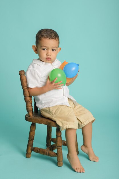 Portrait of smiling little boy dressed in white playing with balloons sitting on wooden chair Blue background