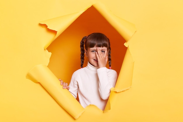 Portrait of smiling little girl with braids wearing white turtleneck posing in torn hole of yellow paper wall peeping spying looking through her fingers at camera