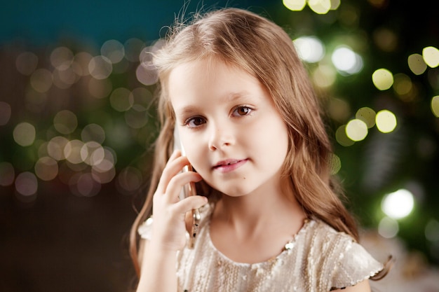 Portrait of a smiling long-haired little girl in dress on  of Christmas lights. Little girl talking on the phone. New Year and Christmas 