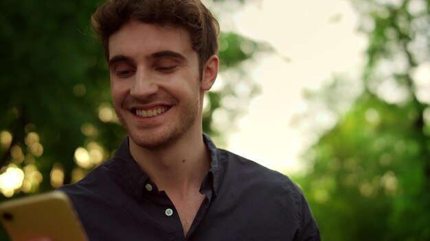 Portrait of smiling man reading message on smartphone outdoors Closeup cheerful guy using mobile phone in park at sunset Young male person sending voice message online on cellphone outside