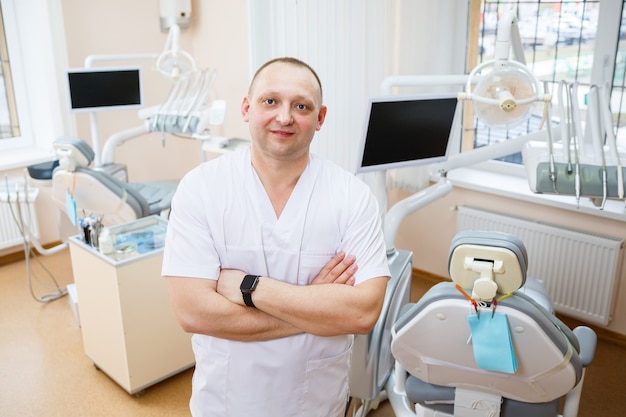 Portrait of smiling man in white doctor robe uniform is standing in office, clinic with chair, instruments. Dentist workplace. Orthodontist is preparing for visit of patient. Dentistry concept.