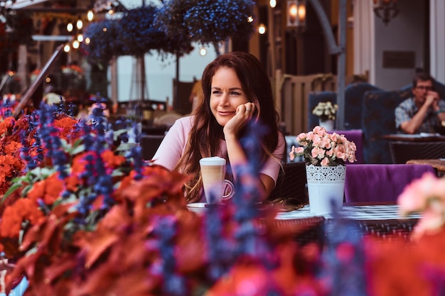 Portrait of a smiling middle age businesswoman with long brown hair wearing a pink dress sitting with a glass of cappuccino at the outdoor cafe.