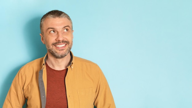 Portrait of smiling middle aged man posing over blue studio background and looking at copy space panorama