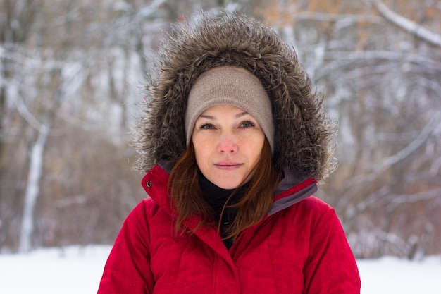 Portrait of smiling middle aged woman with calm look looking at camera in winter park in warm red cloth with hood