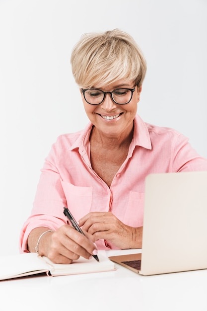 Portrait of smiling middle-aged woman with short blond hair wearing eyeglasses using laptop computer and notepad isolated over white wall