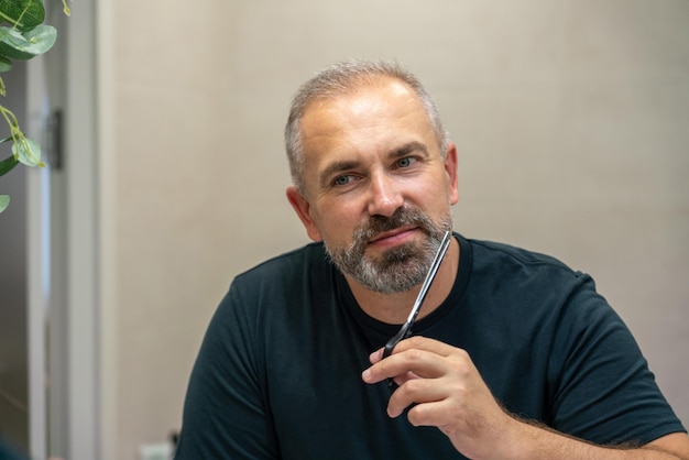 Portrait of smiling middleaged handsome man cutting his beard with scissors selfcare during