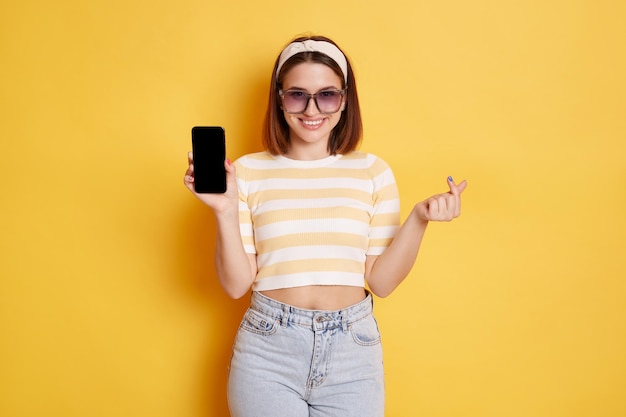 Portrait of smiling optimistic woman wearing striped t shirt and jeans holding smart phone with space for advertisement and showing korean mini love gesture posing isolated over yellow background