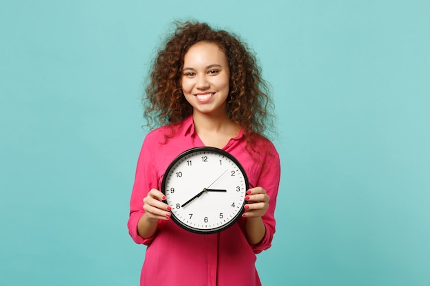 Portrait of smiling pretty african girl in pink casual clothes holding round clock isolated on blue turquoise wall background in studio. People sincere emotions, lifestyle concept. Mock up copy space.