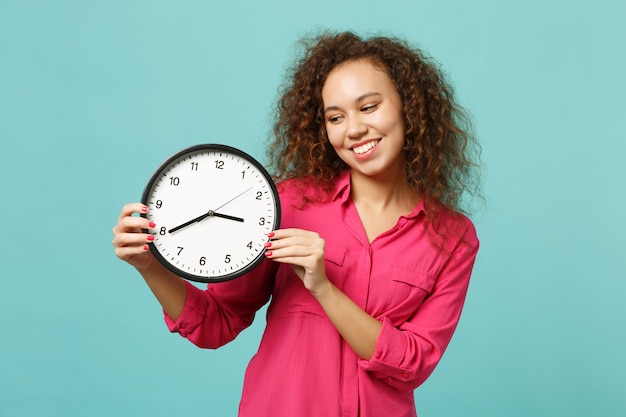 Portrait of smiling pretty african girl in pink casual clothes holding round clock isolated on blue turquoise wall background in studio. People sincere emotions, lifestyle concept. Mock up copy space.