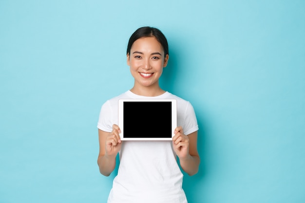 Portrait of smiling pretty asian female showing digital tablet screen with pleased expression, demonstrate shopping site, application for artist, standing light blue wall