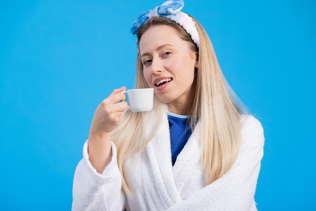 A portrait of a smiling pretty blonde getting up first thing in the morning in a bathrobe drinking a cup of coffee to wake up a good start to the day Blue background in the studio