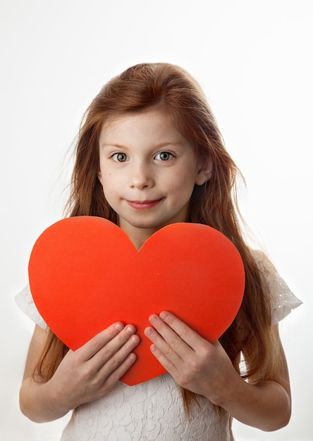 Portrait of smiling red-haired little girl holding big red heart in her hands on white background. Love, Valentines Day, Mothers Day or Fathers Day concept.