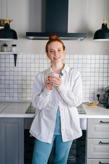 Portrait of smiling redhead woman holding cup of hot beverage in hands on morning at kitchen