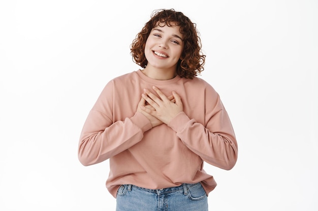 Portrait of smiling romantic girl, feeling touched or heartfelt, holding hands on heart and looking grateful at camera, appreciate something, standing over white background