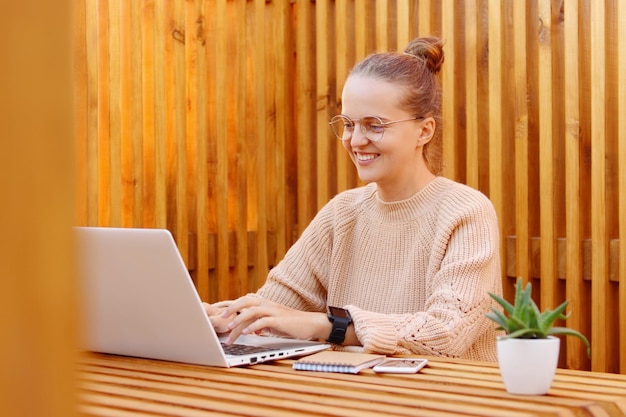 Portrait of smiling satisfied delighted woman wearing beige sweater working on laptop in office against wooden wall typing on notebook with happy facial expression