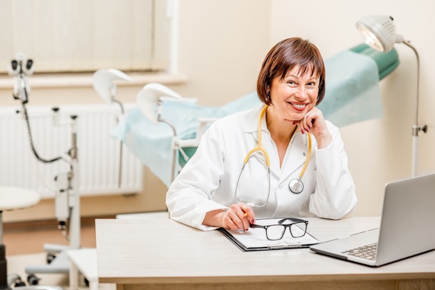 Portrait of a smiling seinor doctor sitting with laptop and documents in the gynecological office