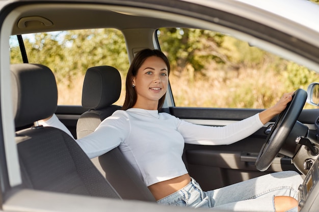 Portrait of smiling self confident dark haired woman wearing white shirt sitting in her car keeps hand on steering wheel looking at camera with smile