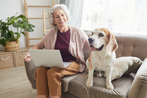 Portrait of smiling senior woman using laptop while sitting with dog on sofa in cozy apartment and smiling