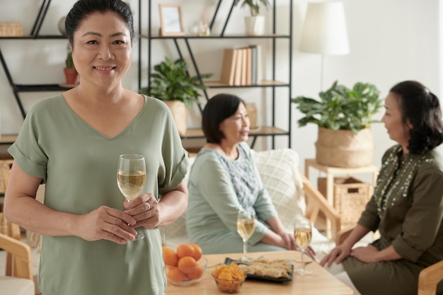 Portrait of smiling senior woman with glass of champagne, her friends chatting in backgrounds