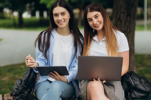 Portrait of smiling student girlfriends using laptop and tablet to study in the park