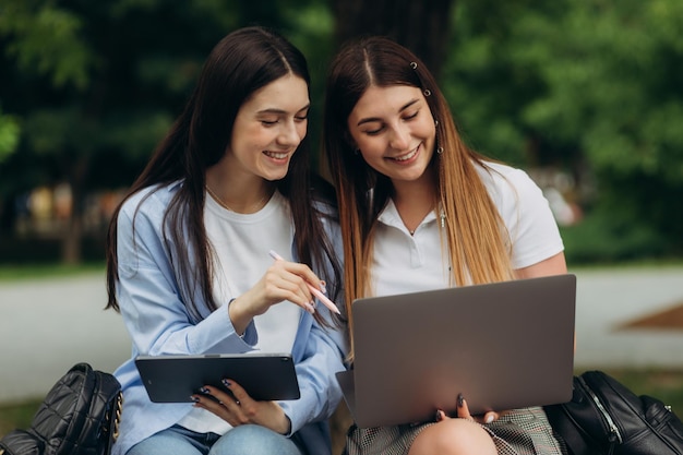 Portrait of smiling student girlfriends using laptop and tablet to study in the park